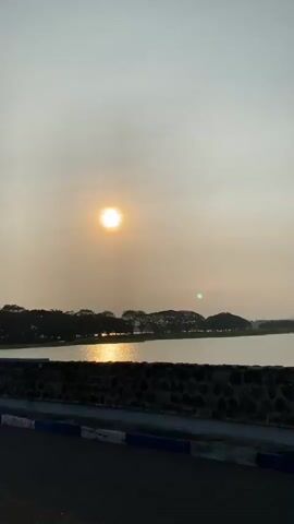 This is a vertical, outdoor video of a sunset over a body of water. The sun is visible as a bright, circular orb in the hazy sky. In the foreground, there is a stone wall with large, metallic letters spelling out "WADUK BENING". Beyond the wall and water, there is a line of trees and some rounded hills. The water reflects the sunlight. The video appears to be a steady shot, with no camera movement.