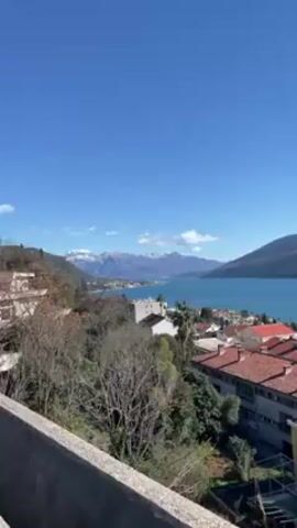 The video presents a landscape view from an elevated position, showing a body of water with mountainous terrain in the background. The water is calm, reflecting a bright blue sky with minimal cloud cover. In the foreground, there are clusters of buildings with red-tiled roofs interspersed with greenery, suggesting a residential area. The angle of the shot is slightly downward, and the camera seems to pan slowly from left to right. The overall scene conveys a sunny, clear day in a coastal town.