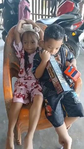 Two young children, a boy and a girl, are sitting closely together on an orange plastic chair. The girl is on the left, wearing a pink pajama set with a cherry pattern and a white headband with fuzzy ears. She has her arms raised above her head. The boy is on the right, wearing a black t-shirt with a graphic design and black shorts with a similar pattern. He is holding a toy that resembles a truck and appears to be chewing on its side. They are sitting outside, with a blurry background showing what looks like a building or structure and two motorcycles parked behind them. The lighting suggests it is daytime.