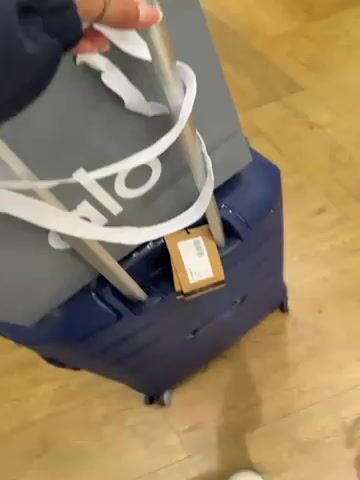 The video shows a person walking and pulling a blue suitcase across a wooden floor. A gray tote bag with white handles and the word "alo" on it sits on top of the suitcase, and a brown tag hangs from the handle. The person wears blue pants and a white shoe. The floor has a light brown, wood-grain appearance. The video is taken from a high angle, focused on the suitcase and the person's legs.