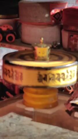 The video shows a close-up of a spinning prayer wheel. The prayer wheel is gold and has Tibetan writing on it. It is sitting on a yellow base which is also spinning. On top of the prayer wheel is a small gold container. In the background, there are stacks of colorful hats.