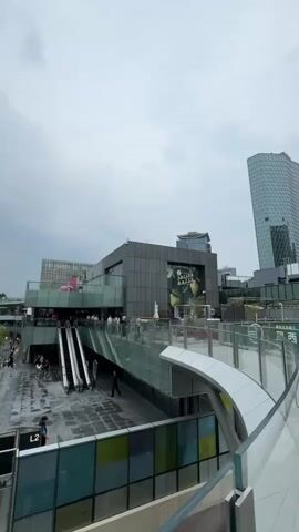 The video shows a busy outdoor plaza area. In the center, there are several escalators leading up to different levels of a modern building complex. People are seen walking on the escalators, in the plaza, and on the elevated walkways. The ground is wet, suggesting recent rain. To the right, a tall, modern glass building stands against a cloudy sky. The architecture of the buildings is contemporary, with glass facades and metal accents. Some shops and advertisements are visible on the building levels. Trees and greenery are also present, adding to the urban landscape. The overall atmosphere appears to be that of a bustling commercial and social hub.