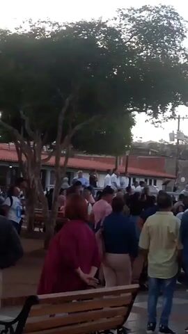 The video shows a crowd of people gathered in an outdoor space. In the background, there are two screens displaying the Venezuelan flag. The crowd is facing the screens, and some individuals appear to be listening to someone speaking from an elevated position near the screens. Trees and buildings surround the gathering, and the sky suggests it's either dusk or dawn.  The scene seems to be a political rally or a similar public gathering.