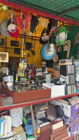 Here's a concise description of the video:

The video shows a cluttered vendor stall, likely at a flea market or antique fair. Various items are displayed including hats, books, CDs, small trinkets, watches displayed in a glass case, old signs (Coca-Cola), and other miscellaneous items. The stall has a red frame, a yellow background, and a green corrugated roof with items hanging from it. Overall, the scene depicts a tightly packed collection of goods for sale.