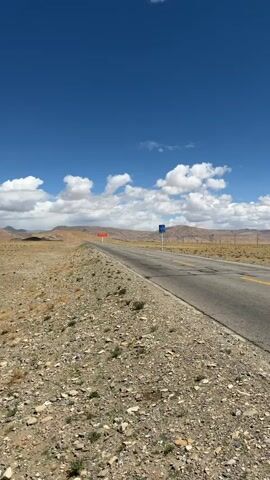 The video captures a wide, expansive view of a remote road stretching through an arid, mountainous landscape under a bright blue sky dotted with fluffy white clouds. A white car, resembling an SUV, drives away from the camera on the right side of the paved road. The terrain surrounding the road is dry, rocky, and sparsely vegetated. In the distance, a red banner-style sign with white Chinese characters is visible across the road, and a blue road sign stands on the right shoulder. The scene depicts a journey through a desolate yet scenic region, likely in Mainpu township, China, given the provided context.