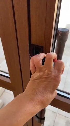 A hand turns a door handle and opens a door, revealing a sunlit patio. The patio has cream-colored tile flooring with decorative accents and is furnished with several chairs and small tables. A white balustrade lines the edge of the patio, beyond which a vast expanse of blue ocean meets a clear blue sky. The scene is serene and appears to be a vacation setting.