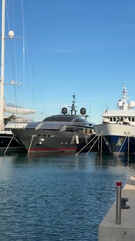 The video pans across a marina on a sunny day, featuring several yachts and sailboats docked close to each other. The water is relatively calm and reflects the blue sky. The first yacht is a sleek, dark grey motor yacht with red accents along its hull. Adjacent to it is a sailboat with a tall mast, dark blue hull, and white upper deck. Further along is a large, white motor yacht, partially visible, named "MELLON". There are no visible people or activities taking place in the immediate frame, creating a serene and upscale maritime scene.