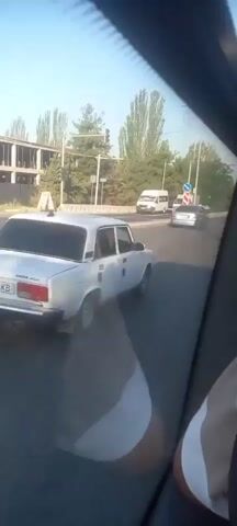 A white car drives down a road, emitting a large cloud of black smoke from its exhaust. The camera is inside another vehicle, looking out of the window at the car. The scene is outdoors with trees and buildings visible in the background. The sky is clear and the sun is shining.