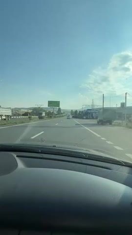 The video is taken from the interior of a car, looking forward through the windshield. It shows a highway with multiple lanes of traffic moving under a clear blue sky with scattered clouds. On the left side of the highway, there is a green billboard with text and graphics. On the right side of the highway, there are several businesses, including what appears to be a gas station with pumps and a large truck parked nearby. Power lines and electrical towers are visible along the side of the road. The car appears to be traveling at a moderate speed. The overall environment suggests a sunny day on a rural or semi-rural highway.