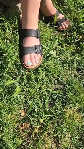 The video shows a woman with tattoos wearing sandals, a white t-shirt, and black shorts. She is sitting on a stone wall outdoors, lighting and smoking a cigarette. The background includes a green field and mountains.