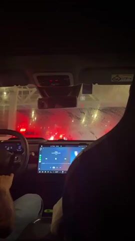 This is a POV video from inside a car at night. The camera is positioned behind two people. In the foreground, a person's hands are visible on the steering wheel and in their lap. They are wearing a blue denim jacket. In the background, another person's head and shoulder are visible, and they are looking out the windshield. The car's infotainment screen is displaying a music player interface. The windshield is wet, with water streaks visible, suggesting it is raining or has recently rained. Outside the car, red lights from traffic or signs are visible through the windshield, reflecting off the wet surface. The overall environment appears to be a street or roadway, possibly in a city, with some kind of structure or overpass visible above the traffic lights. The lighting is dark, with the primary light sources being the car's interior lights, the infotainment screen, and the external red lights.