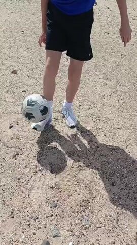 A person is shown from the waist down standing on a sandy, rocky surface. They are wearing black shorts, white socks, and white athletic shoes. The person is juggling a soccer ball, which is white with black and green accents. The ball is suspended in the air, at knee level, with the person's left foot positioned beneath it, ready to make contact. A shadow of the person and the ball is cast on the ground to the right. The video shows a continuous action of the person controlling the ball.
