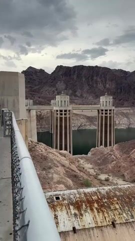 The video shows a sweeping panoramic view of a dam and its surrounding landscape. The dam, made of concrete, is partially visible in the foreground, with a rusty, weathered surface. Beyond the dam, a body of water, likely a reservoir, is visible. The water level appears to be significantly low, with exposed rock formations and debris lining the edges. Two tall, rectangular towers with pointed tops stand in the middle ground, emerging from the water. In the background, rugged, mountainous terrain with dark, rocky slopes dominates the horizon. The sky is overcast with heavy, gray clouds. The scene is a stark depiction of a dry environment with evident signs of drought.