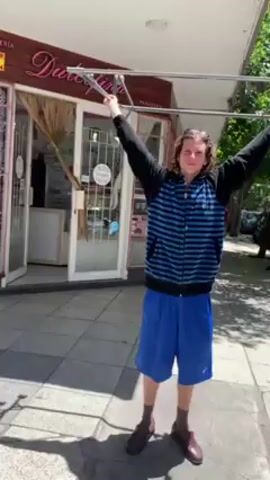 The video depicts a person holding a metallic structure above their head on a city sidewalk. The person, with long brown hair, is wearing sunglasses, a blue-striped hooded jacket, blue shorts, brown socks, and brown shoes. They are standing in front of a storefront with the sign "Dulcepa" visible. The sidewalk is paved with gray tiles and extends along a street with other storefronts and trees lining the side. Several people are visible walking down the street in the background. The lighting indicates a sunny day. The person holding the structure appears to be smiling and turns to look down the street.