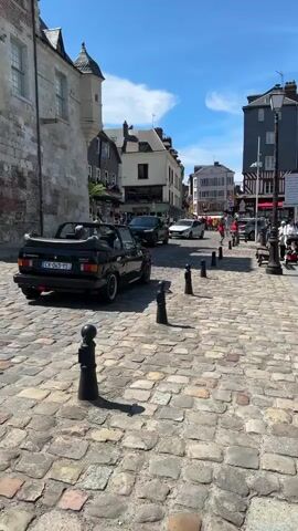 The video shows a sunny day in a European town with cobblestone streets. A black convertible is parked on the left side of the street. In the background, there are several people walking and cars driving. The buildings are old and made of stone and brick. The sky is blue with a few clouds.