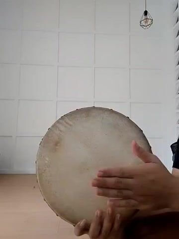 A person's hands are seen playing a drum in a room with white paneled walls and a wooden floor. A light fixture hangs from the ceiling. The person is using their hands to strike the drum, creating a rhythmic sound. The drum is round and made of a light-colored material, possibly animal hide. The person's hands are positioned in different ways to create different sounds on the drum.