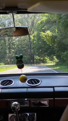 The video shows the interior of a car, looking out the windshield. The car is driving on a winding road through a forest. The road is paved and has a guardrail on the side. There are several road signs indicating sharp turns. The dashboard of the car is visible, with a chrome steering wheel that has a chain wrapped around it. There are also some dashboard ornaments hanging from the rearview mirror. The driver's arm, with tattoos and a bracelet, is visible on the steering wheel. The trees are lush and green, suggesting it is summertime. Sunlight is filtering through the trees, creating dappled light on the road. The overall impression is a scenic drive through a wooded area.