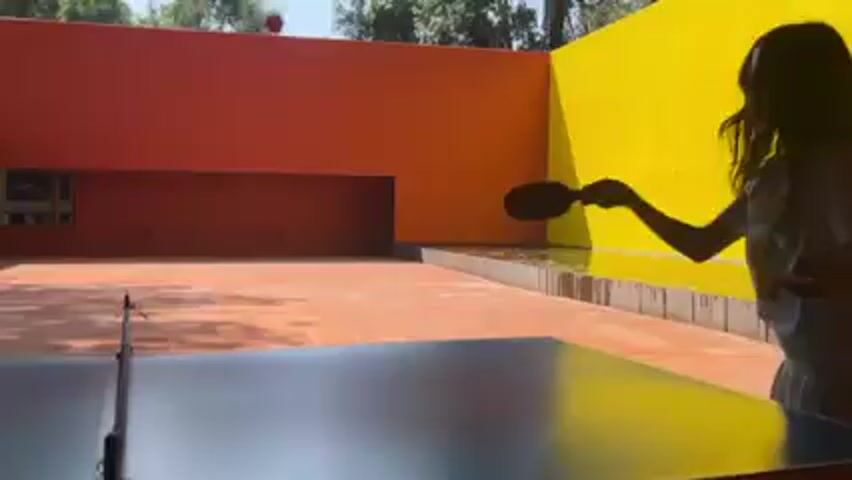 The video shows an outdoor ping pong match in progress. The camera's point of view is looking across the table. An orange ping pong ball is being hit back and forth. A woman is playing. The playing area is a tiled patio with a large orange building in the background and a covered area to the left, with wooden posts, a thatched roof, and seating. There are trees in the background. The flooring below the table is tiled, and there are some twigs and leaves on the ground.