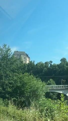 This video shows a castle on a hill under a clear blue sky. The castle is made of light-colored stone and has a pitched roof. It appears to be surrounded by trees and vegetation. There is also a bridge visible in the foreground, and some power lines are strung across the sky. The video seems to be filmed from a moving vehicle, as the perspective shifts slightly throughout.