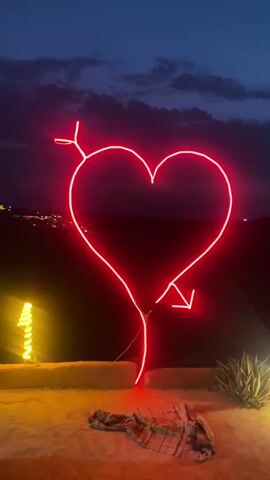 A neon red heart-shaped sign, pierced by an arrow, is illuminated against a dark, cloudy sky. The sign stands on a rocky, outdoor surface. In the foreground, a blanket or cloth is spread on the ground. To the left, a yellow light source glows. Distant lights suggest a town or settlement in the background.