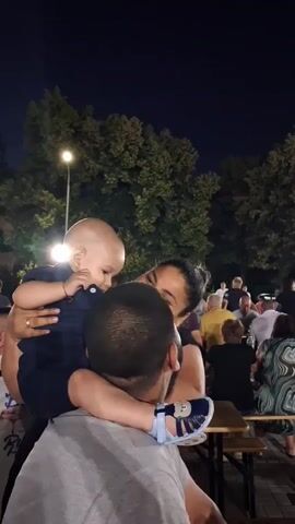 The video captures a tender moment between a family at an outdoor gathering in Malomir, Bulgaria. A man, viewed from behind, is holding a baby on his shoulders. The baby, dressed in a dark blue outfit, is being embraced by a woman who is looking down at the child. The scene appears to be an evening event, as indicated by the dark sky and the illumination from streetlights and ambient lighting in the background.

In the background, numerous people are seated at picnic tables, suggesting a festive atmosphere, perhaps a community event or a celebration. Trees line the perimeter of the area, and a lamppost with a bright light is visible. The overall mood is warm and familial, with the focus on the intimate interaction between the parents and their child. There is no visible text within the frame. The weather appears to be pleasant, suitable for an outdoor evening gathering. The presence of Bulgarian flags in the far left of the frame confirms the location context.