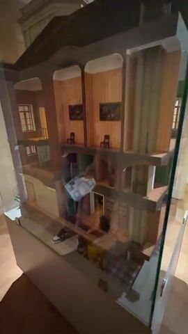 A dollhouse is viewed from the exterior, with glass covering the front. The dollhouse is multi-story and appears to be a replica of a historical building. The camera pans across the interior rooms, revealing furniture, miniature paintings on the walls, and architectural details such as spiral staircases and doorways. A person's reflection is visible in the glass, and at one point, their hand is seen reaching into the dollhouse. The rooms are furnished in a period style, with wood paneling, chandeliers, and fireplaces. The lighting is dim, creating a moody atmosphere.