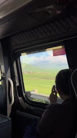 The media file shows a view from inside a bus, looking out the window at a scenic landscape. The primary subject is a person sitting on the bus, seen from behind, with their hand near their face, possibly holding a phone to take a picture or for another purpose. The person is wearing a dark-colored garment.

The window reveals a bright, sunny day with a vast expanse of lush green fields. In the distance, a village with houses is visible nestled at the base of rolling hills. Beyond the hills, majestic mountains with snow-capped peaks rise against a clear blue sky with scattered white clouds. The overall scene suggests a journey through a rural, mountainous region.

A notable detail is a red and white sticker on the window with text, which reads "EMERGENCY EXIT" in English, and some text in Georgian script above it. The time of day appears to be daytime, given the bright sunlight and the clarity of the landscape. The weather seems pleasant. The location context provided is Tsilkar, Armenia, which aligns with the mountainous terrain and village scenery. The mood evoked is one of peaceful travel and appreciation for natural beauty.
