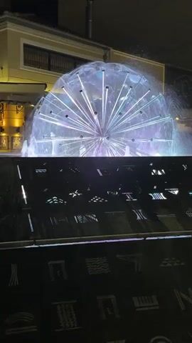 The video showcases a vibrant spherical water fountain illuminated at night. The fountain is designed with numerous jets that spray water outwards from a central point, forming a large, translucent sphere. Internal lights within the fountain cycle through various hues, predominantly white, blue, and purple, casting a mesmerizing glow and reflecting off the moving water.

Below the main fountain structure, a dark, perforated panel is visible. This panel features an array of geometric and abstract cut-out patterns, such as wavy lines, triangles, stripes, and arch-like shapes. Colored light shines through these openings, highlighting the intricate designs.

In the background, part of a building with windows and a chimney on its roof can be seen, partially illuminated. The overall scene is set at night, with the artificial lights of the fountain and panel providing the main illumination. The fountain is actively spraying water throughout the clip. There are no people visible, and no text is present in the frame. The location is Karditsa, Greece.