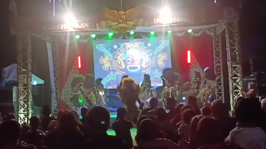 A group of dancers in elaborate costumes are performing on a stage. The stage is decorated with red curtains and illuminated by bright lights. A large screen behind the performers displays a colorful image of a mask. A crowd of people are watching the performance from the audience area in front of the stage. The event appears to be taking place outdoors at night. The dancers are moving in unison, and their costumes are detailed and striking. The overall atmosphere seems festive and celebratory.