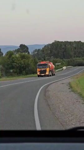 The video shows a view from inside a vehicle, looking out at a road. A yellow truck with a red tank is driving on the road. The road curves to the right and goes over a small bridge. There are trees and fields on either side of the road, and mountains in the distance. A sign with blue lettering that says "р. БЕЛОКУРИХА" is visible on the right side of the road. The sky is overcast and appears to be dusk. Another car is seen approaching in the distance.