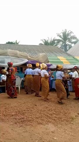 The video shows a group of women gathered outdoors, likely at a celebration or event. Most of the women are dressed in similar attire: a white top, a yellow and brown patterned skirt, and a matching headwrap. Some wear different clothing, including a red skirt with a pattern and a patterned dress. They stand on a reddish-brown dirt ground, and some appear to be dancing or moving to the music. A drummer sits on a white chair, playing a drum set. In the background, there is a large tent or awning with a multi-colored roof. Trees and buildings are visible in the far background.