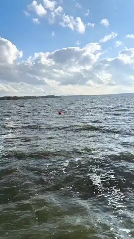 The video shows a wide shot of a large body of water under a partly cloudy sky. The water is choppy, with visible waves and foam. In the distance, a faint treeline is visible on the horizon. There are several red buoys scattered across the water. The sky is blue with scattered white clouds, and sunlight is visible. The video appears to be shot from a moving boat, as indicated by the slight camera shake and the wake behind the viewer. There are no people or distinct actions visible in the video, only the environment and the buoys.