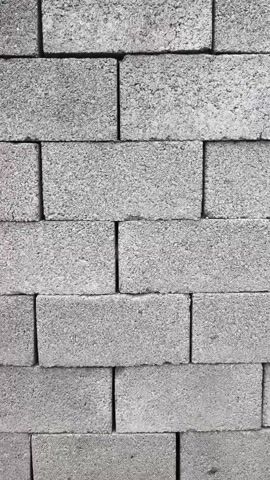 A close-up, vertical shot shows a stack of grey concrete blocks. The blocks are arranged in a staggered pattern, with each layer offset from the one below it. The texture of the blocks is rough and porous, typical of concrete. The lighting is even, with no harsh shadows, suggesting an overcast sky or artificial lighting. The camera appears to be static. The video is short, approximately five seconds long, and there is no discernible audio. The focus is sharp, capturing the granular detail of the concrete.