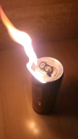 The video shows a can of liquid on fire. The can is dark in color and features a silver pull-top lid with two openings. Flames are emerging from these openings, extending upwards and to the left. The can is positioned on a flat, reflective surface, possibly a wooden table or countertop. The background is dimly lit, suggesting a dark or indoor setting.