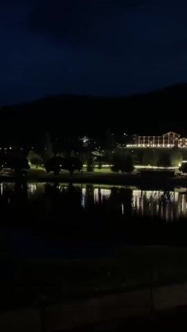 The video shows a nighttime scene with a body of water in the foreground and a large building complex on a hill in the background. The building is illuminated with numerous lights, creating a warm glow. Trees are scattered around the building and along the shoreline. The dark sky is visible above the hill. The lights from the building reflect on the surface of the water. The scene appears serene and peaceful.