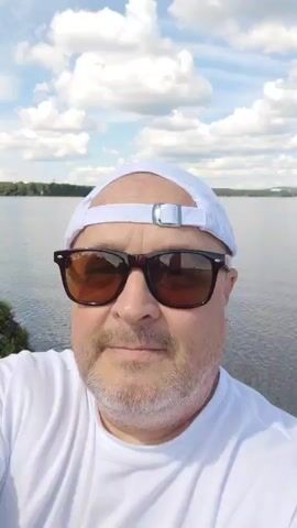 A medium shot captures a Caucasian man wearing a white t-shirt, a white baseball cap, and dark sunglasses, with a slight beard. He is smiling and looking directly at the camera. Behind him, a body of water stretches to the horizon, with a faint treeline and distant structures visible. The sky above is blue with scattered white clouds. The image is a still frame, suggesting a photograph or a paused video.