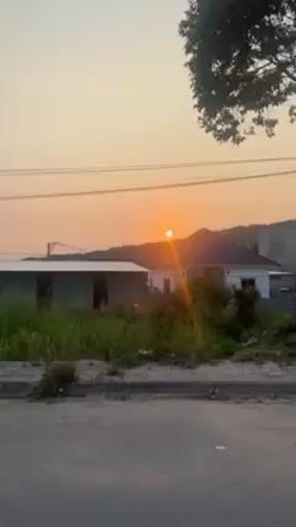 The video depicts a vibrant sunset, the sun appearing as a bright orange orb low on the horizon, casting a warm glow over the scene. The sun is partially obscured by a distant mountain range. Silhouetted against the sky are several buildings with corrugated metal roofs and utility poles crisscrossed by numerous power lines. A tree with dense foliage stands in the foreground. The camera pans upward slightly, enhancing the view of the sunset and the power lines. The video appears to be shot from a moving vehicle along a paved road. The overall atmosphere is serene, accentuated by the sunset's beauty.