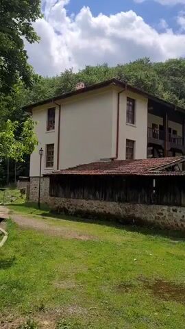 The video presents an outdoor scene featuring an architectural complex, likely a monastery, set against a backdrop of a dense green forest and a partly cloudy blue sky. The camera executes a smooth horizontal pan from left to right. Initially, a two-story building with cream-colored walls, dark wooden balconies on the upper floor, brown window frames, and a red tile roof is visible. A dirt path and green grass are in the foreground, along with a low stone wall topped with horizontal wooden planks. As the camera pans right, piles of cut logs appear on the grass in front of the stone wall. Subsequently, a stone church building with multiple domes, characterized by yellow-orange and white tops and red tile sections, comes into view. This church features stone walls and arched windows. A dark paved path is visible in the foreground, leading towards a dark gate within the stone wall. No individuals are present in the scene.