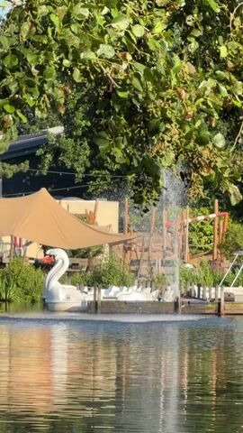 A tranquil lake scene unfolds under a canopy of green leaves. In the foreground, the water reflects the dappled sunlight. On the far bank, a white swan-shaped paddleboat rests by a dock. Behind it, a series of white paddleboats are lined up. A tall fountain sprays water in the center. Further back, a tan awning provides shade over a seating area. Behind the awning, a wooden playground structure with red accents is visible. The environment appears to be a recreational area, possibly a park or resort, with lush vegetation surrounding the lake.