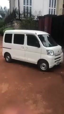 The video begins with a close-up of a white van parked on a red dirt surface. The camera then pans right, revealing a street with two individuals walking away from the camera. The street is unpaved and bordered by concrete walls on either side. The individuals are walking away, making it difficult to discern details of their clothing or appearance. The environment appears to be a residential area with visible buildings and greenery in the background.