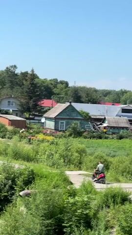 A still image shows a rural landscape under a clear blue sky. In the foreground, lush green bushes and trees dominate the frame. A dirt path winds through the greenery. Several houses are visible in the middle ground, with one prominent dark wooden house with a sloping roof and a white-trimmed window. To its right, a lighter-colored house with a red roof is partially obscured by trees. Another house, painted green and white, stands further to the right. A person is riding a red motorized vehicle, possibly a scooter or ATV, down the dirt path, moving away from the viewer. Flowerbeds with colorful blooms are visible near one of the houses. The overall impression is a serene, sunlit summer day in a village or countryside setting.