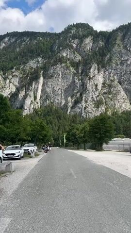 The video shows a road leading towards a large, rocky mountain. Several cars are parked on the left side of the road, and a few people are walking on the road, heading away from the camera. Trees and foliage surround the base of the mountain, and the sky is partially cloudy. On the right side of the road, there are some greenhouses or similar structures. The video is shot from a slightly elevated perspective, looking forward down the road.