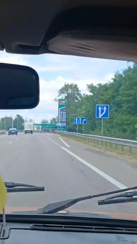 The video shows a view from inside a vehicle, looking out the windshield towards a highway and a gas station. The road has white dashed lines marking the lanes and solid white lines on the shoulder. There are traffic signs indicating lane usage. A gas station with green canopies is visible in the distance, along with a tall sign displaying fuel prices. Trees line the right side of the road. A rearview mirror is visible on the left side of the windshield, and a portion of a Ukrainian flag is attached to the windshield wiper on the lower left. Other vehicles, including a white truck and a blue car, are seen on the highway. The sky is cloudy.