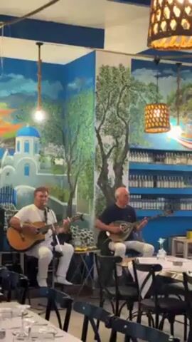 Two middle-aged men are playing instruments in a restaurant. One man is playing the guitar and the other is playing a bouzouki. The men are both dressed in white pants. The restaurant is decorated with a mural of a Greek island scene. The tables are set with white tablecloths and glasses. The room is lit by hanging lamps.