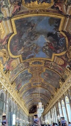 The video shows a low-angle view of a grand, ornate ceiling with elaborate paintings and gilded decorations. The ceiling is arched and stretches the length of what appears to be a long hall. Figures in the paintings are depicted in a classical style, with dramatic poses and vibrant colors, against a backdrop of sky and clouds. The gold trim is intricate, with repeating patterns and architectural details. A large, crystal chandelier hangs from the ceiling, adding to the opulence of the space. In the foreground, at the bottom of the frame, are several people holding up their smartphones, presumably to take pictures or videos of the ceiling. They are in a crowded indoor space, as suggested by the presence of multiple individuals and the background architecture visible through the people's camera lenses. The lighting suggests an indoor environment, possibly a museum or a historical palace, illuminated by natural light filtering in from unseen windows and the chandelier.