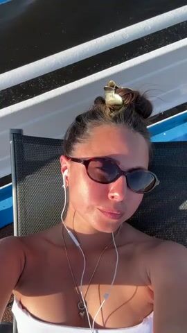 A woman with her hair in two buns, wearing sunglasses and earbuds, lies back in a lounge chair. She is outdoors, with a body of water and striped structures visible in the background. Sunlight casts shadows on her face and chest.