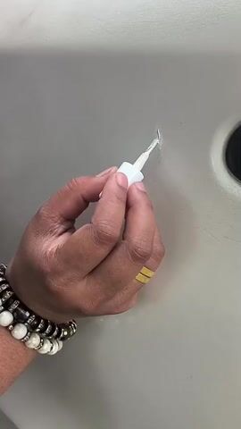 A close-up video shows a person's hand applying a white liquid from a small applicator bottle onto a scratch on a smooth, light-colored surface. The hand wears multiple bracelets, featuring white, black, and pearl beads, as well as a gold-colored ring on the ring finger. The applicator has a slender white handle and a small brush tip. The white substance is carefully dabbed onto the linear scratch, which appears on a surface that is likely light gray or beige. After the application, the hand and applicator move away, revealing the scratch partially covered by the white liquid. A dark, circular object is partially visible on the right side of the frame. The activity suggests a repair or cosmetic touch-up being performed on the surface. While the location context of Lambarotunong, Indonesia is provided, there are no specific visual cues within the video to confirm this setting.