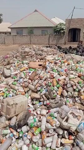 The video shows a large pile of discarded plastic containers and other waste materials in a sandy, open area. The pile is composed primarily of white and colored plastic bottles and canisters, along with some larger plastic containers and darker, unidentifiable debris at the top. The background features a low brick wall and a few buildings with simple architectural designs, typical of residential areas. The sky appears overcast, suggesting a daytime setting. There are no identifiable people or interactions visible in the frame.