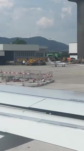 The video shows an airport tarmac on a sunny day. In the foreground, the edge of a white airplane wing is visible. In the middle ground, a small white propeller plane is taxiing on the tarmac. Behind the propeller plane, there is a tall, cylindrical control tower. Further in the background, there are several airport buildings, yellow service vehicles, and green trees. The sky is partly cloudy. The camera pans slightly to the right throughout the video.