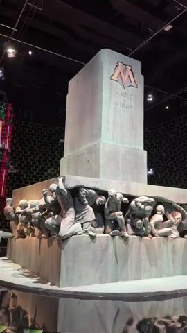 The video shows a set from the Harry Potter films, specifically the Ministry of Magic atrium.  In the foreground is the imposing "Magic is Might" statue, depicting numerous figures struggling under the weight of a large rectangular slab with the Ministry of Magic's emblem. The statue is a dull grey color. Behind the statue, the setting is a dark, tiled room. In the background, a mural depicting a wizard hangs on a wall behind a tiered set, while to the right is a red and green building with multiple windows.  Some people can be seen standing on the set. The lighting in the room is dark, but spotlights illuminate the set.